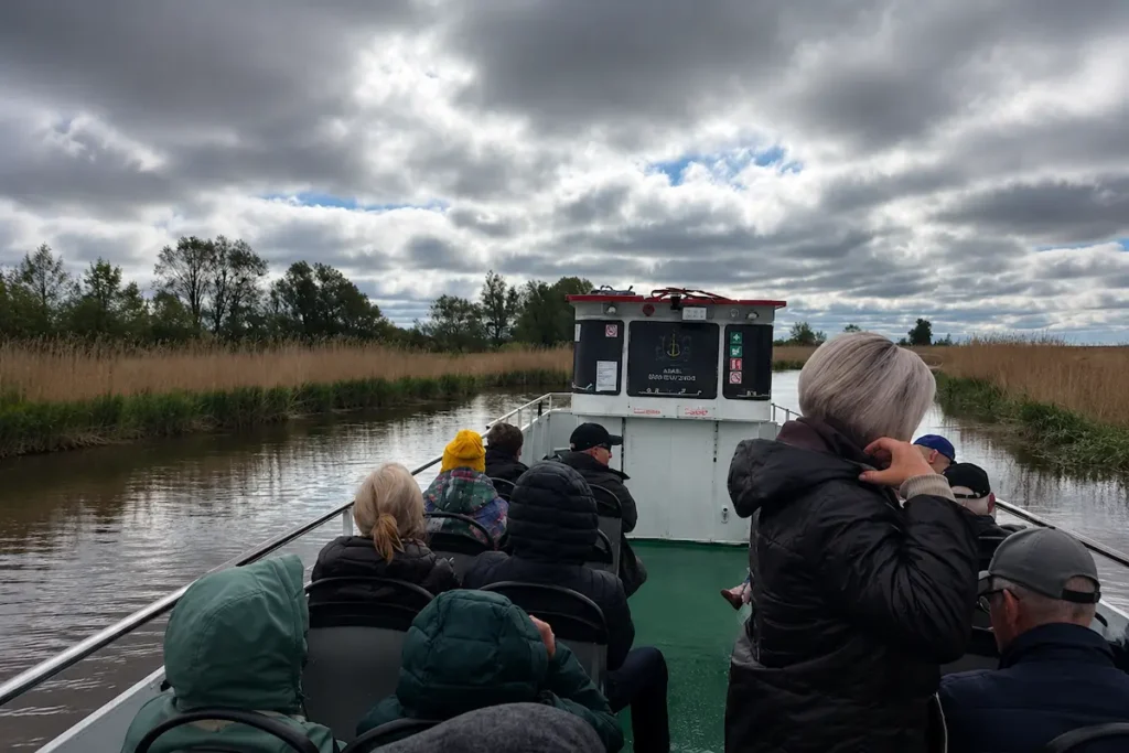 Eine Gruppe von Passagieren unterschiedlichen Alters sitzt auf Holzbänken auf dem Außendeck eines Ausflugsschiffes unter einem weißen Sonnensegel, im Hintergrund eine ruhige Wasserstraße und grüne Bäume.