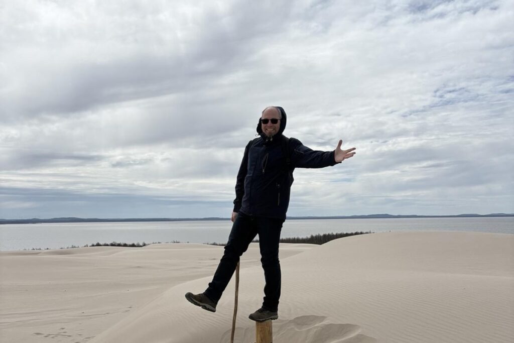 Ein dynamisches Foto des Reiseleiters Wojciech, der voller Energie auf dem Kamm der Lontzker Düne (Wydma Łącka) im Slowinzischen Nationalpark posiert. Im Hintergrund der helle Sand der Wanderdüne unter einem weiten Himmel – ein Symbol für Lebensfreude und Leidenschaft bei der Arbeit.