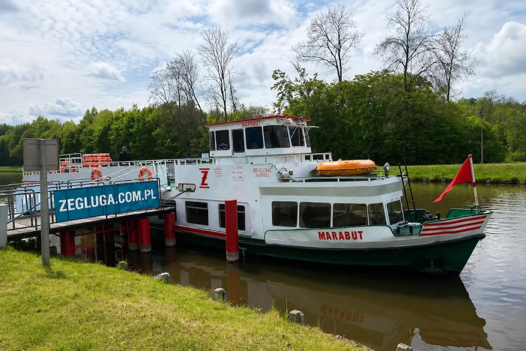 Ein weiß-grünes Ausflugsschiff namens Marabut, angelegt an einem Holzsteg auf dem Oberländischen Kanal in Polen, im Hintergrund dichter grüner Wald und ein leicht bewölkter blauer Himmel.